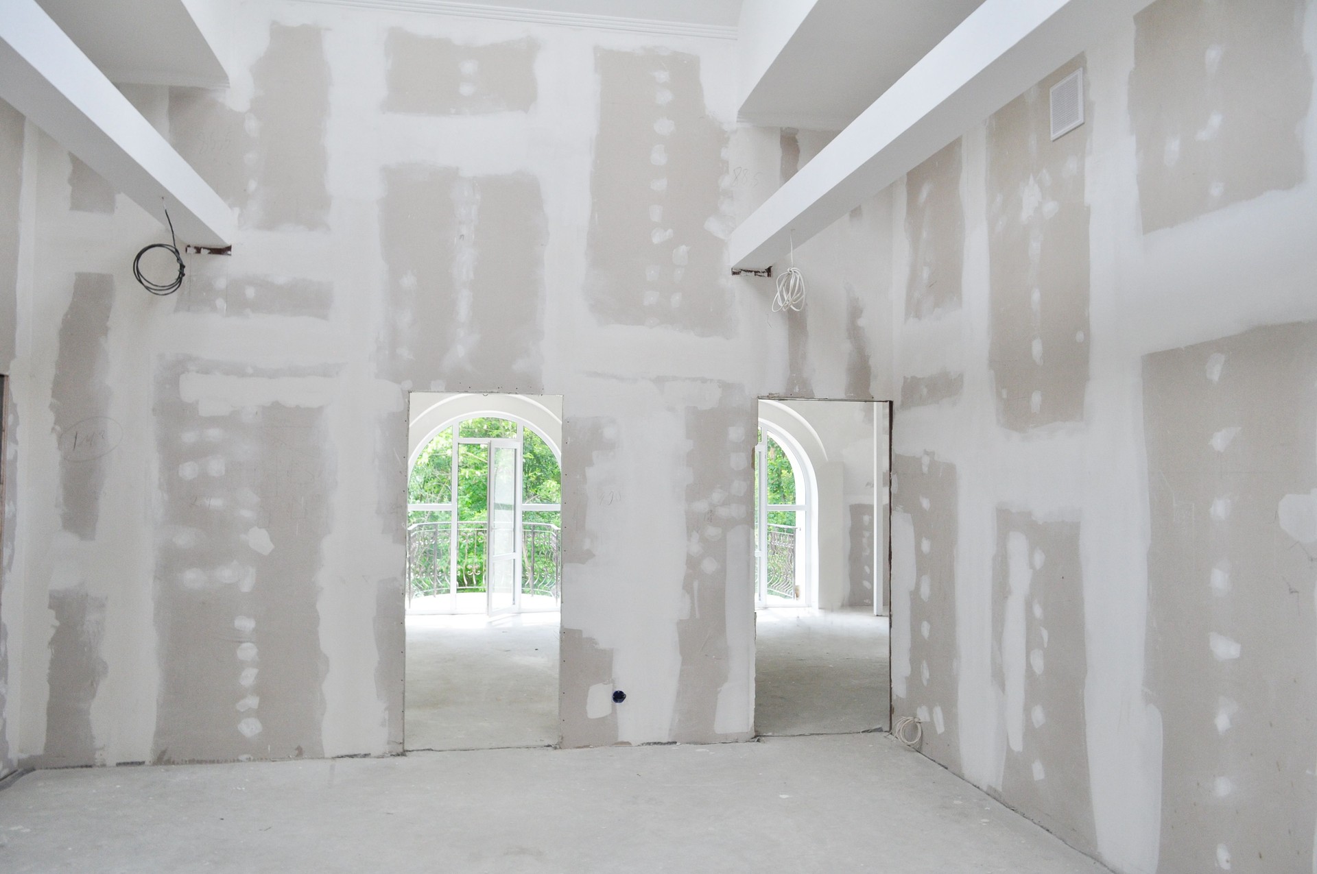 Interior view of a residential building under construction, showing newly installed drywall with taped and mudded seams. The room features high ceilings with exposed beams and arched doorways leading to bright windows that open to a balcony. The smooth drywall finish and clean layout highlight the precision and quality of the interior finishing work in progress. “New drywall installation and finishing in a residential construction project — showcasing smooth seams, high ceilings, and precise interior craftsmanship by a professional construction company.” drywall installation, interior finishing, residential construction, drywall taping and mudding, home renovation, professional construction company, drywall contractor, new home build, interior framing and drywall. Rejuvenation Renovations.