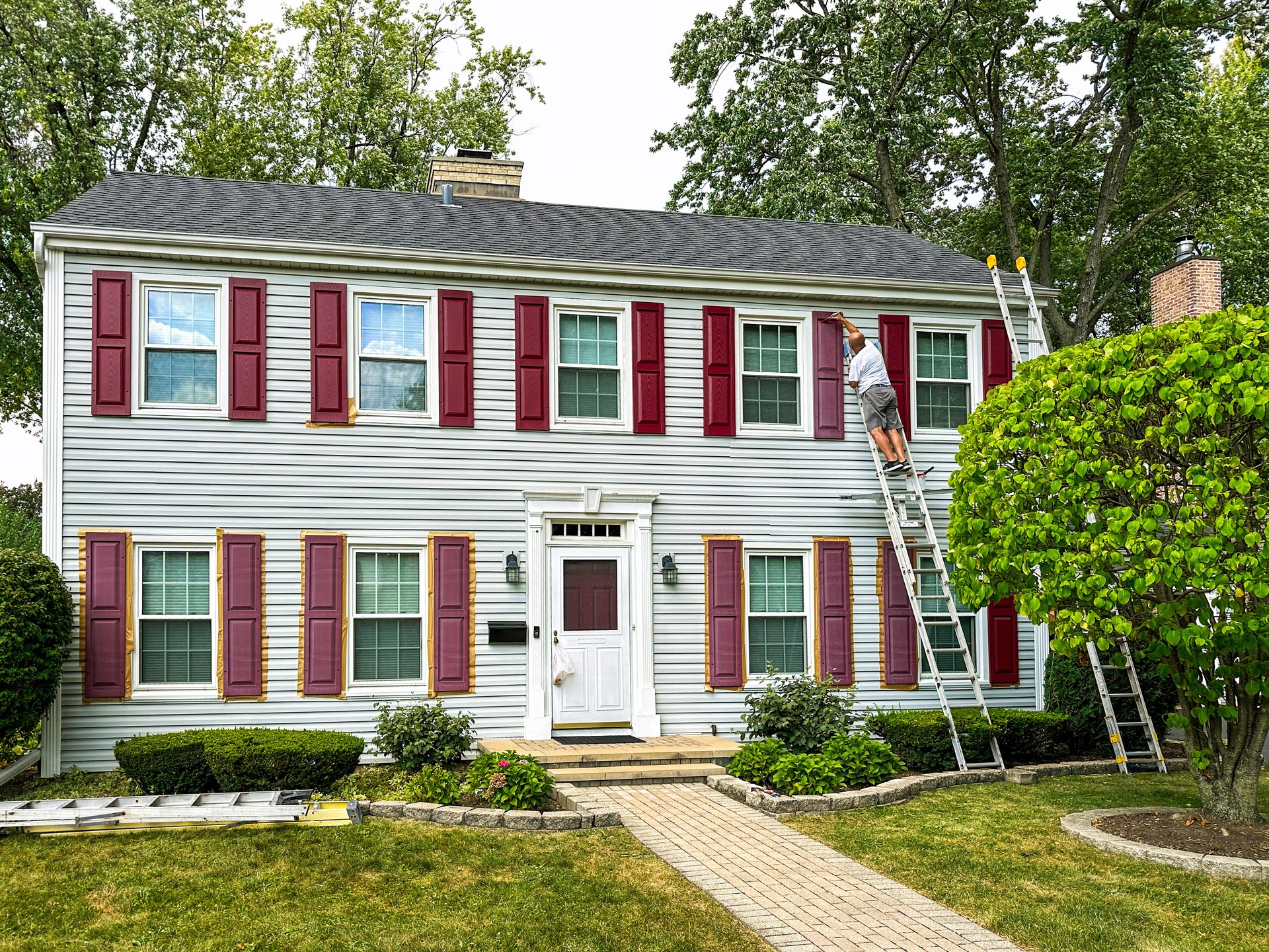 Man at the top of a ladder painting window shutters of a two story white home. Rejuvenation Renovations.