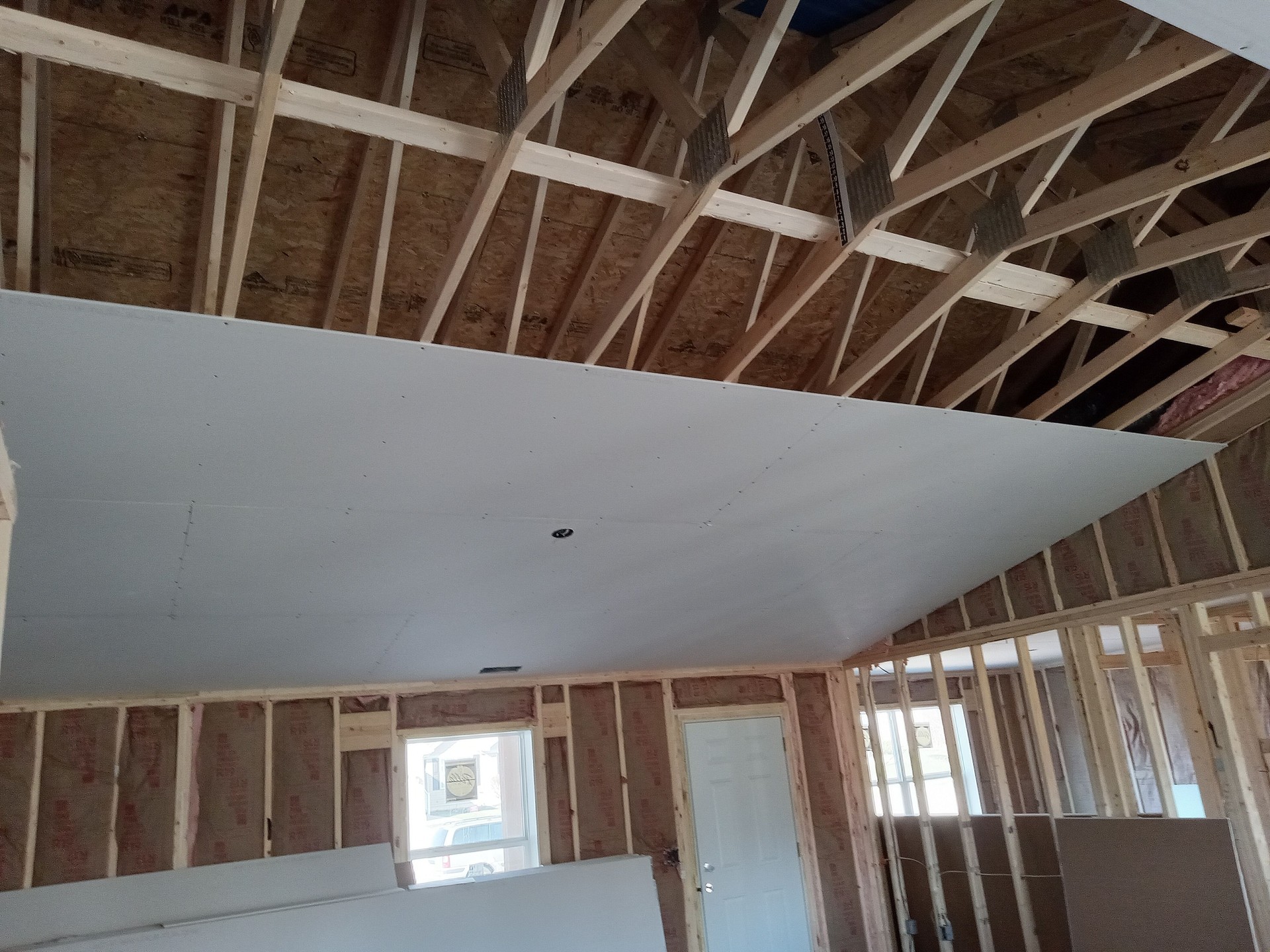 Interior of a residential construction project featuring drywall installation on a vaulted ceiling. The exposed wood framing, roof trusses, and wall insulation highlight the structural phase of the build. A door and window allow natural light to illuminate the workspace. The image demonstrates professional craftsmanship, attention to detail, and quality construction practices during the home-building process. “Residential construction in progress — expert drywall installation and precision framing by a professional construction company, showcasing quality workmanship and structural integrity.” residential construction, drywall installation, framing and insulation, vaulted ceiling build, professional construction company, home building services, quality craftsmanship, new home interior, Rejuvenation Renovations